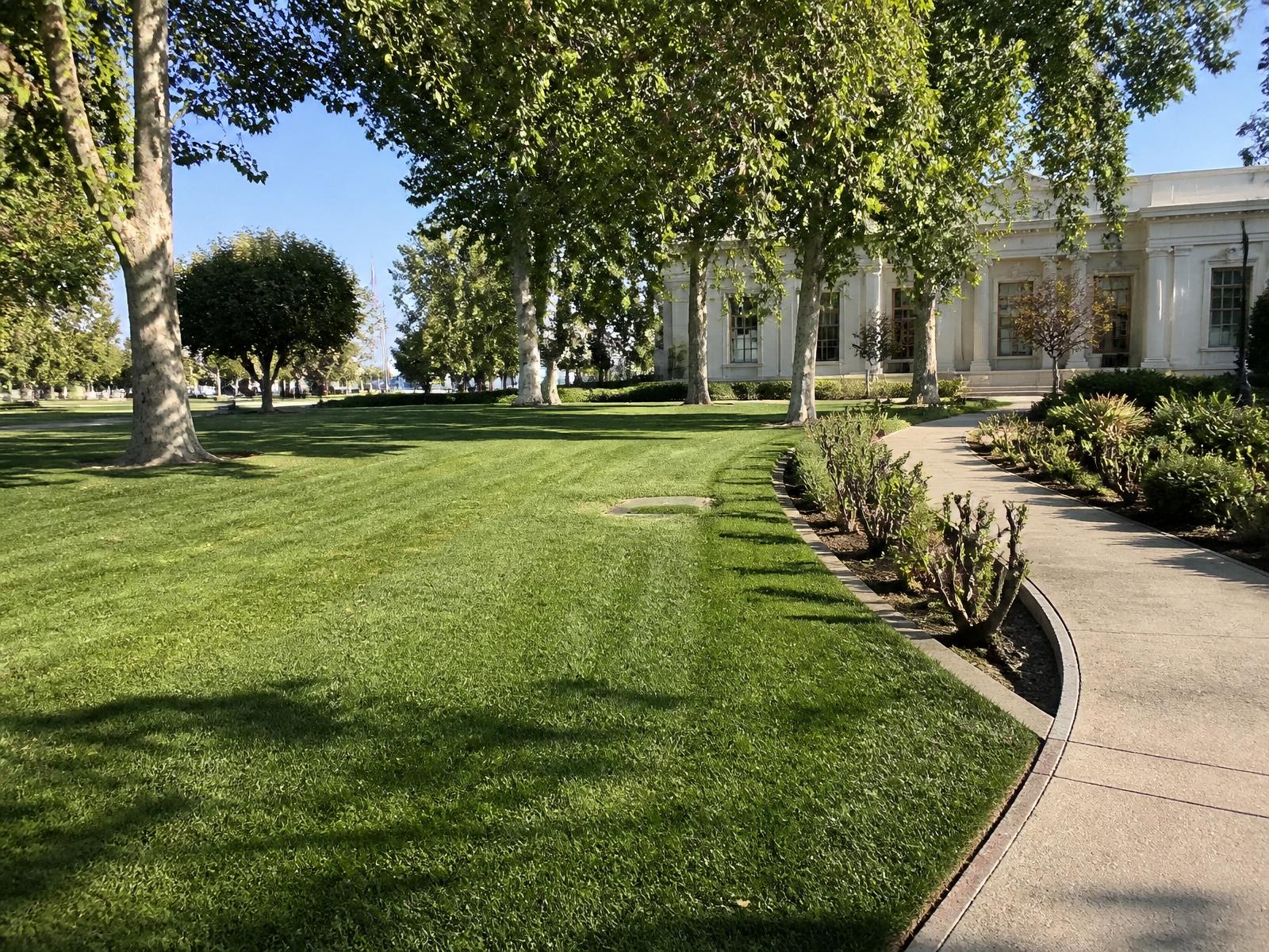 Freshly mowed civic lawn with mature shade trees, curved walkway and pruned planting beds at an institutional building, representative imagery